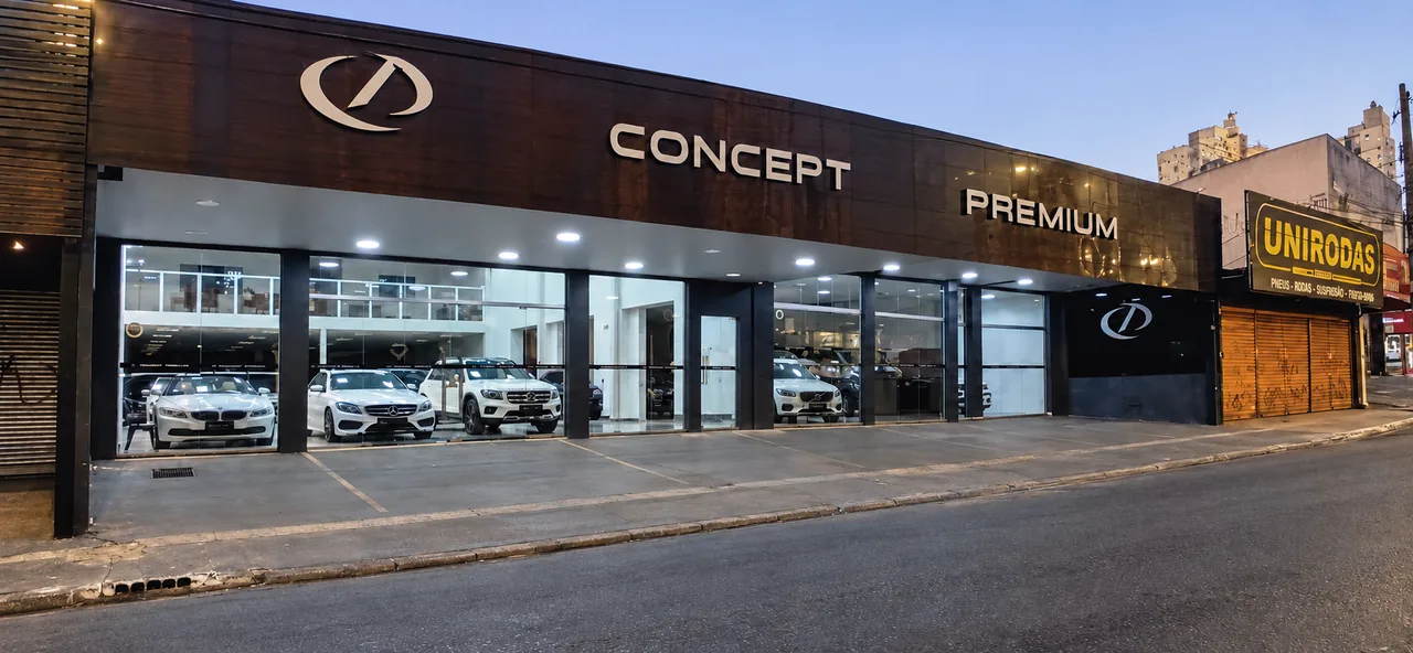 Fachada da Concept Premium na Rua S-1 ao entardecer, com o símbolo e o letreiro em metal sobre a madeira e o showroom de vidro com os carros